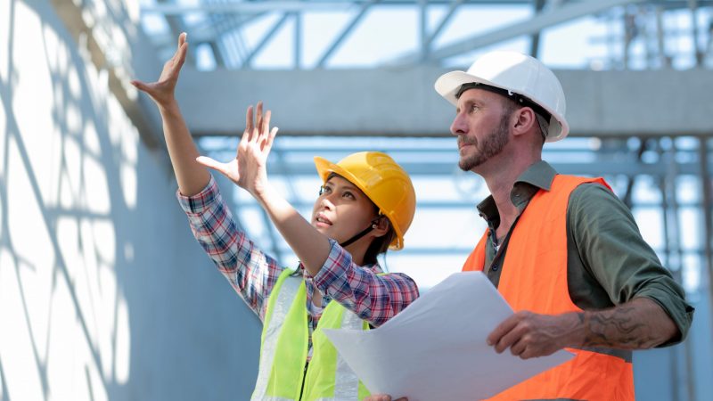 Asian female architect explaining design to white male engineer for building decoration, young building owner listens to report on construction results, side shot.